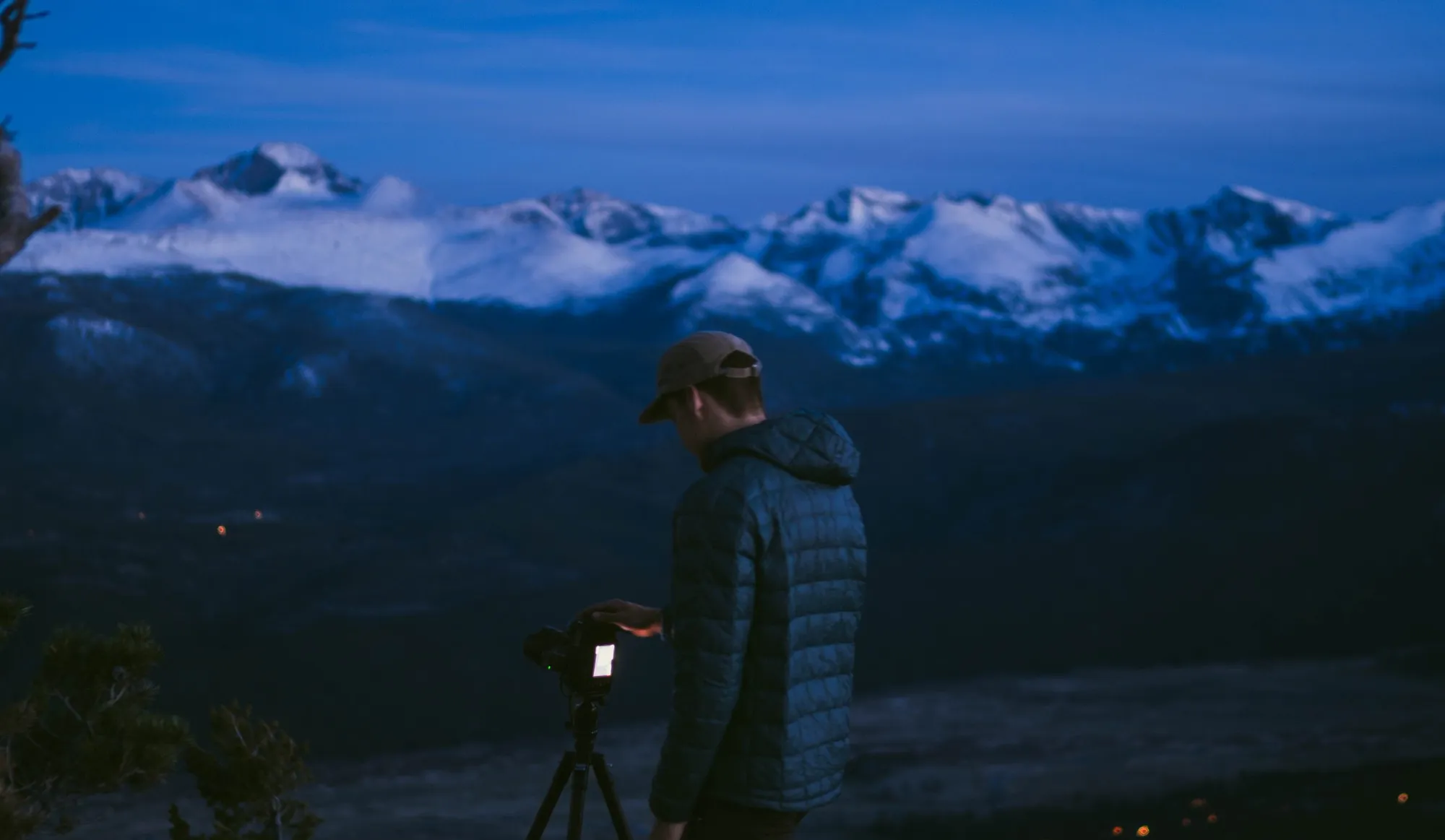 Bryan Vossen adjusting a camera on a tripod at blue hour, snow-covered peaks behind him