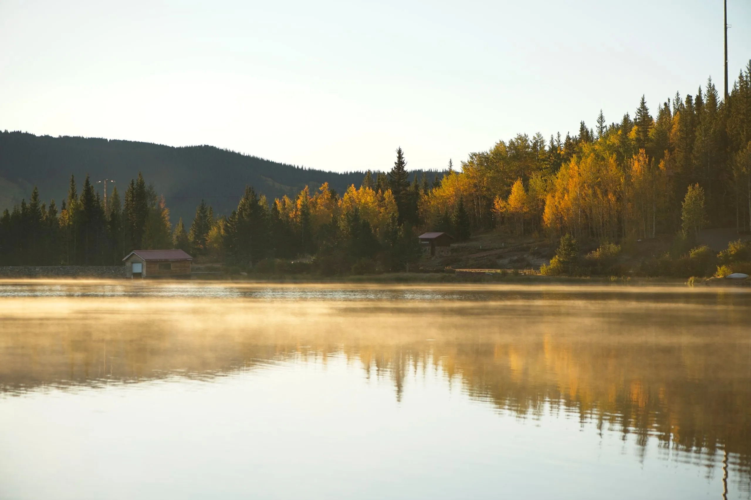 Golden aspens and dark evergreens reflected in a calm lake, mist rising off the water and small cabins on the far shore