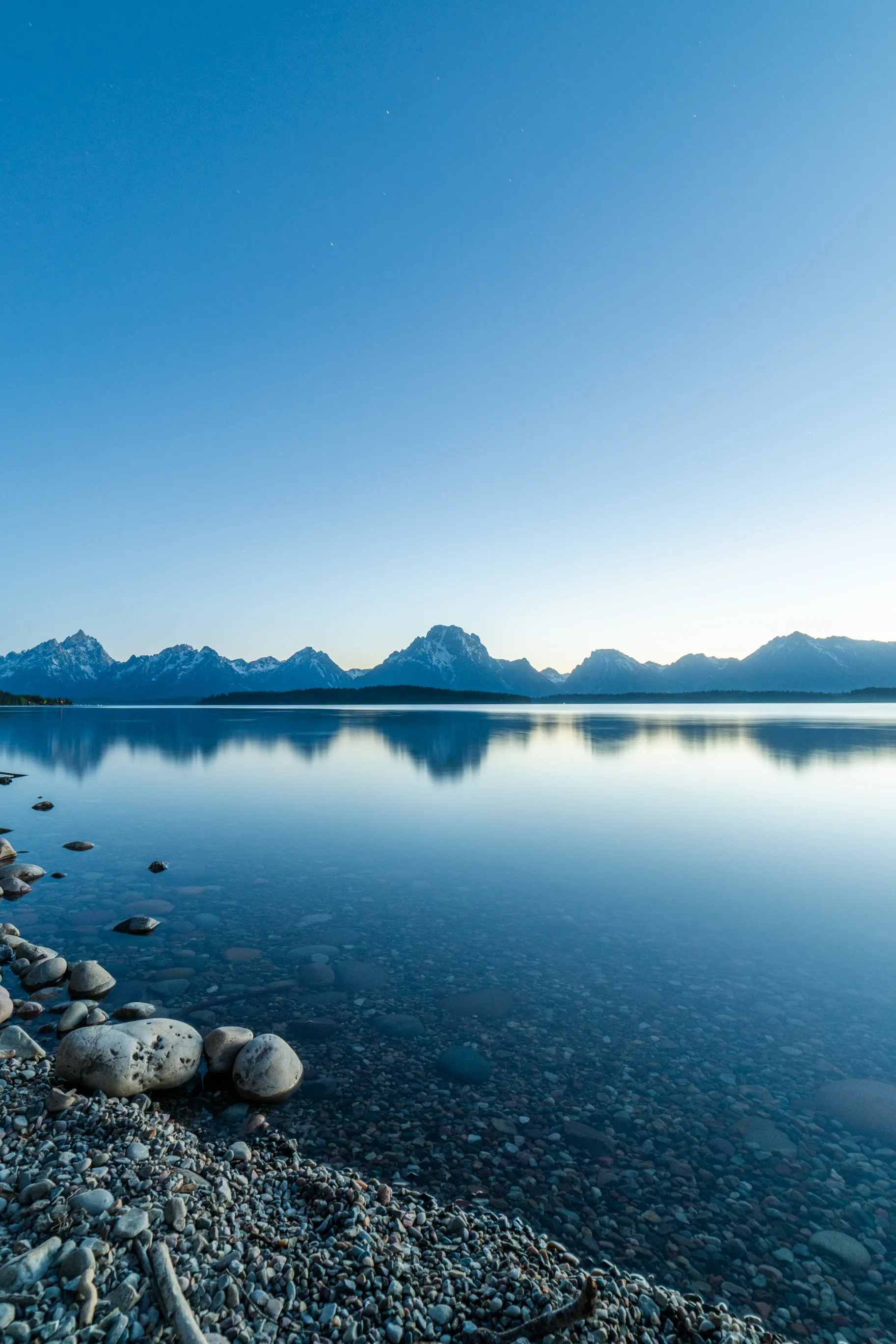 A glassy lake at blue hour, rounded stones in the foreground and the Teton Range on the horizon