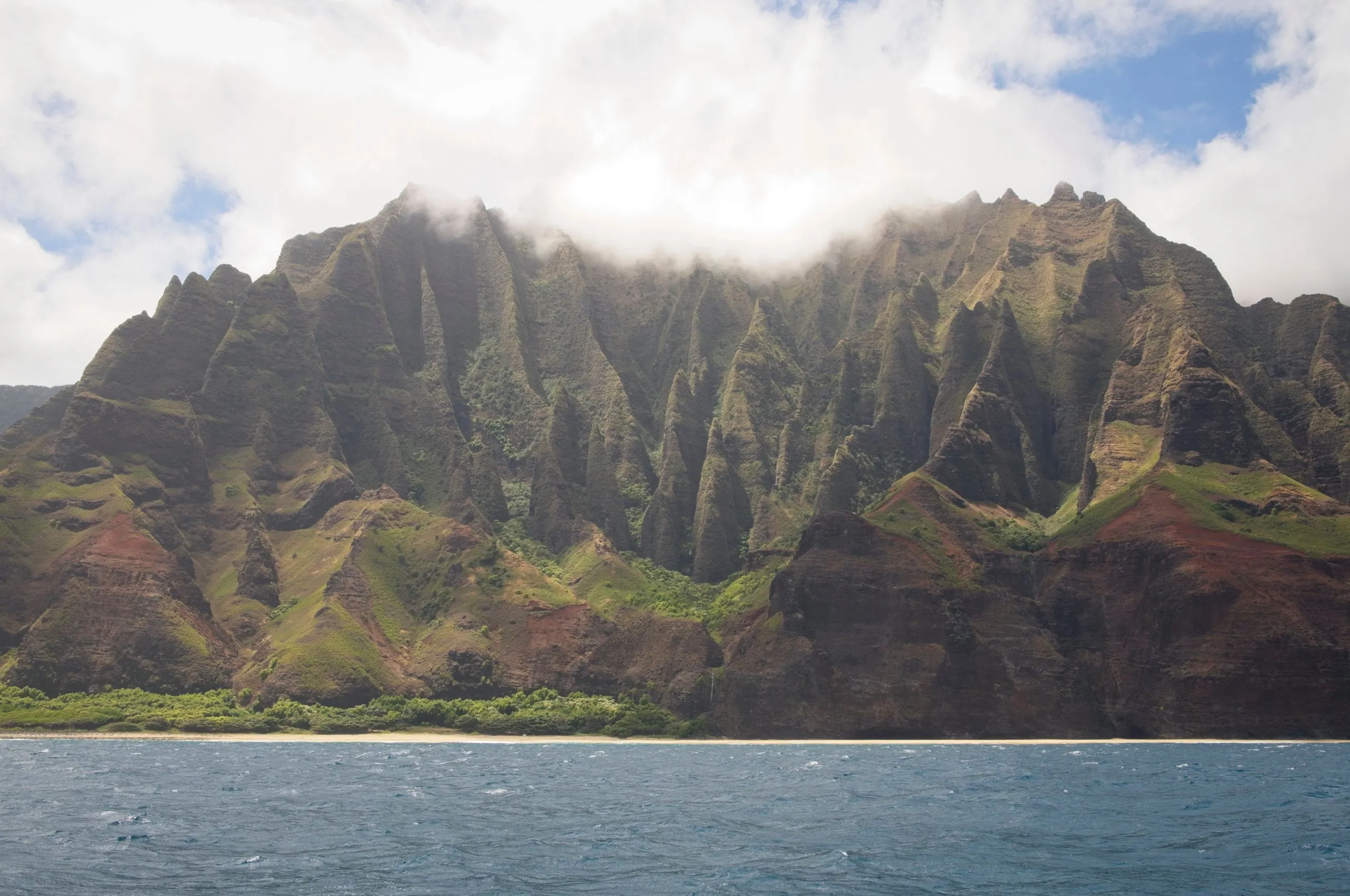 Steep, fluted green ridges of the Nā Pali Coast rising straight from the ocean, their peaks wrapped in cloud