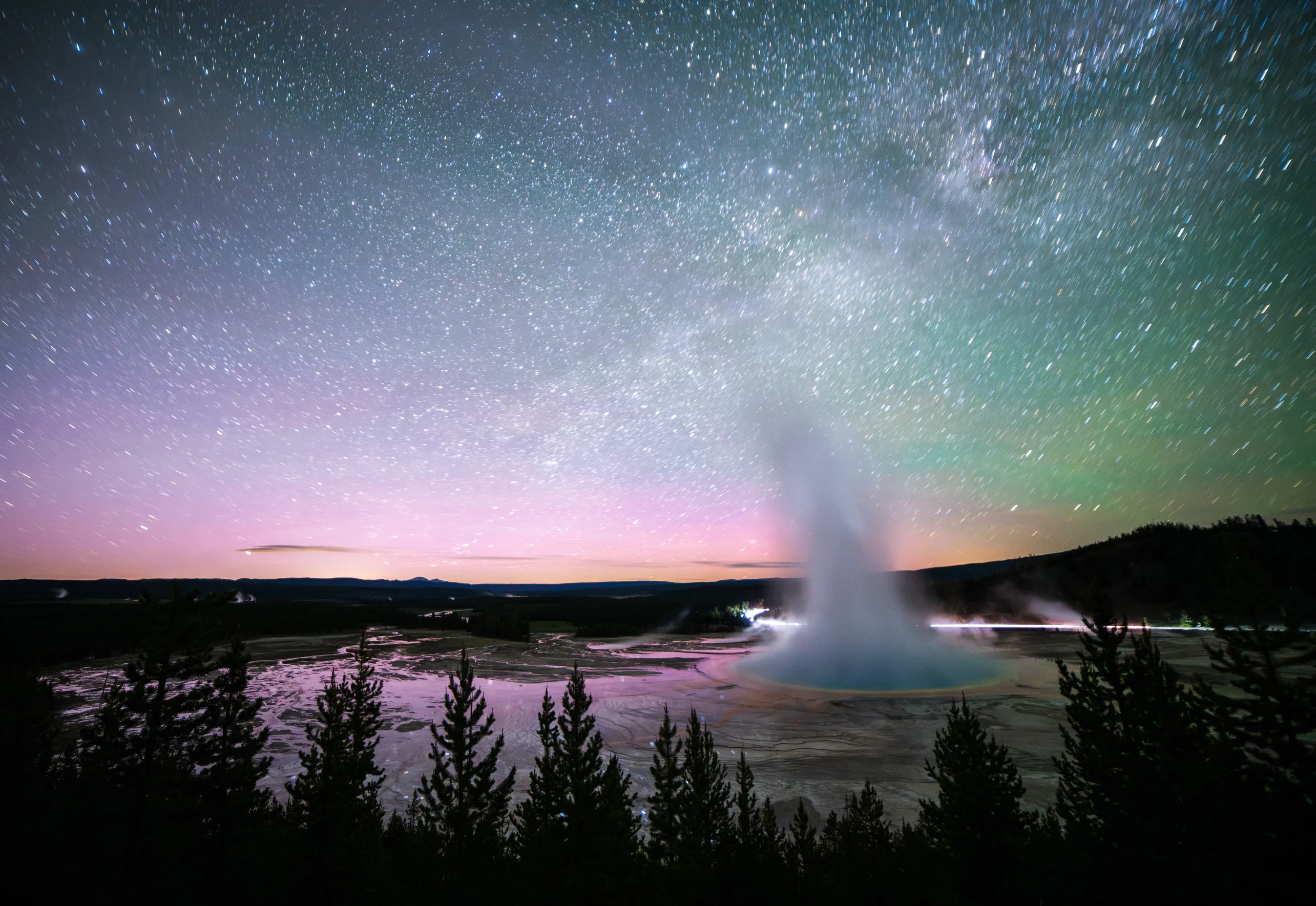 A geyser erupting at night beneath a starry sky with the Milky Way and a pink glow on the horizon, dark pines in the foreground