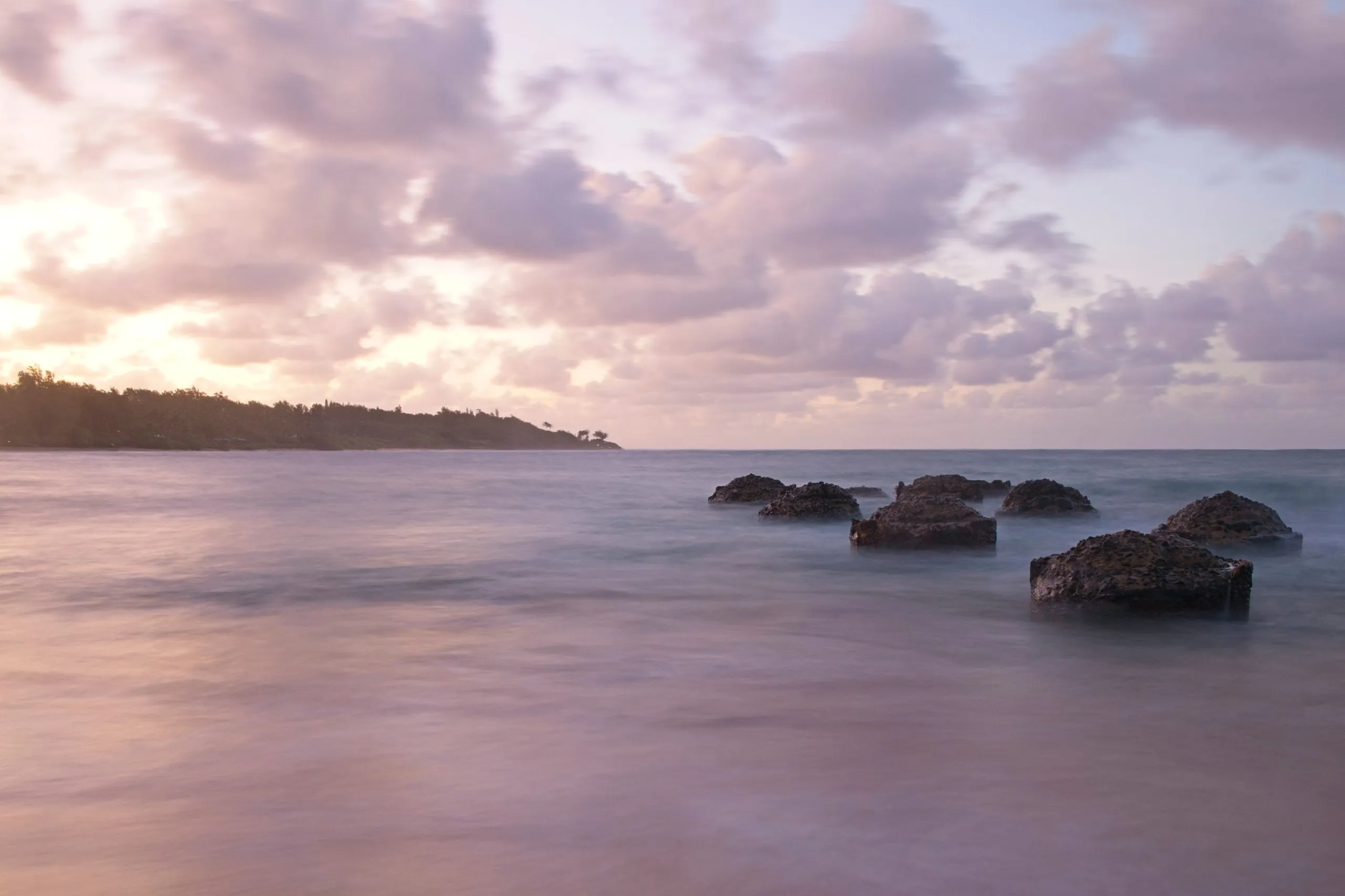 Silky water flowing around dark lava rocks beneath pink and lavender clouds at dusk, with a wooded point in the distance
