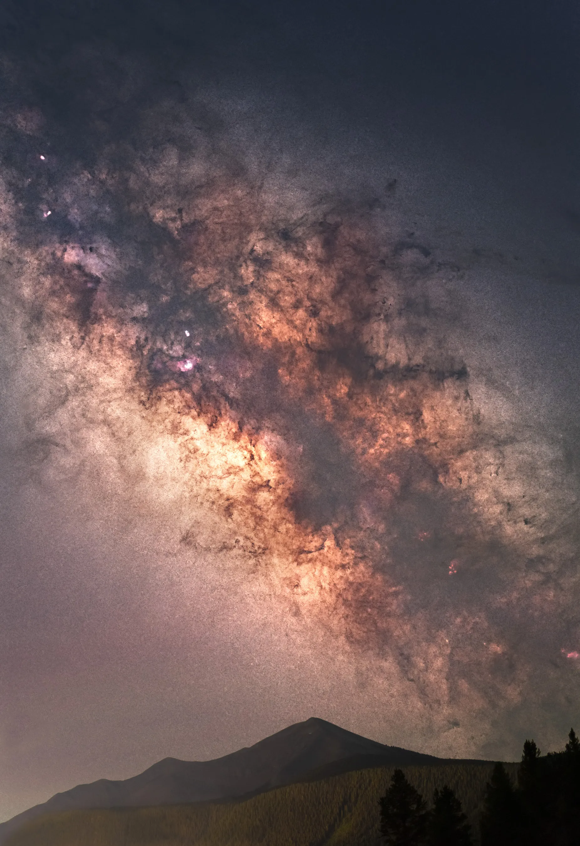 The bright core of the Milky Way filling the sky above a lone mountain peak and silhouetted trees