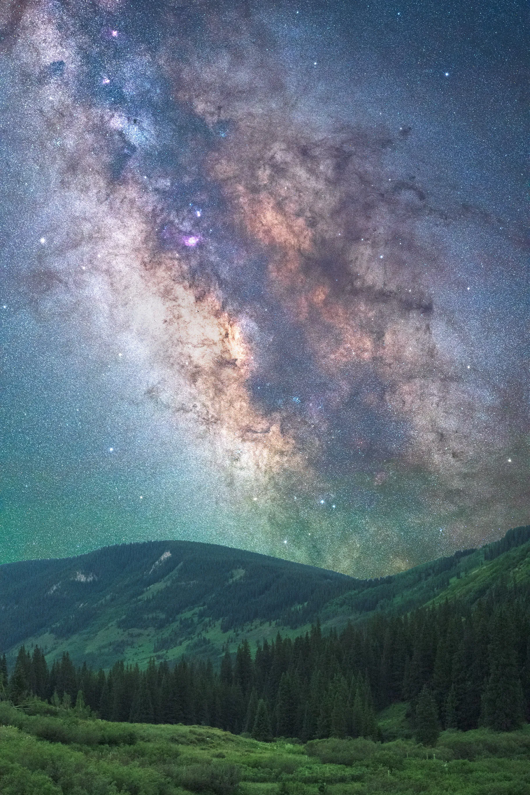 The Milky Way stretching over a green mountain valley and dark evergreen forest