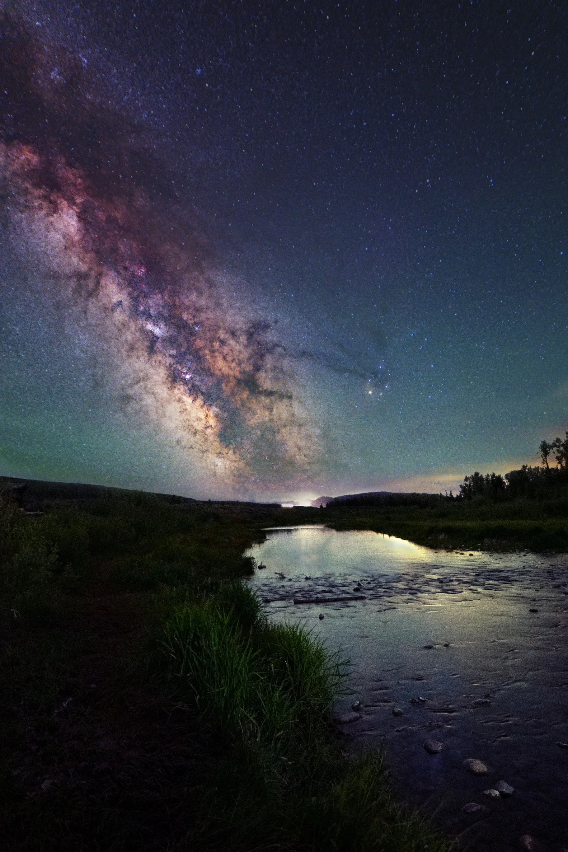 The Milky Way arching over a shallow river winding between dark grassy banks