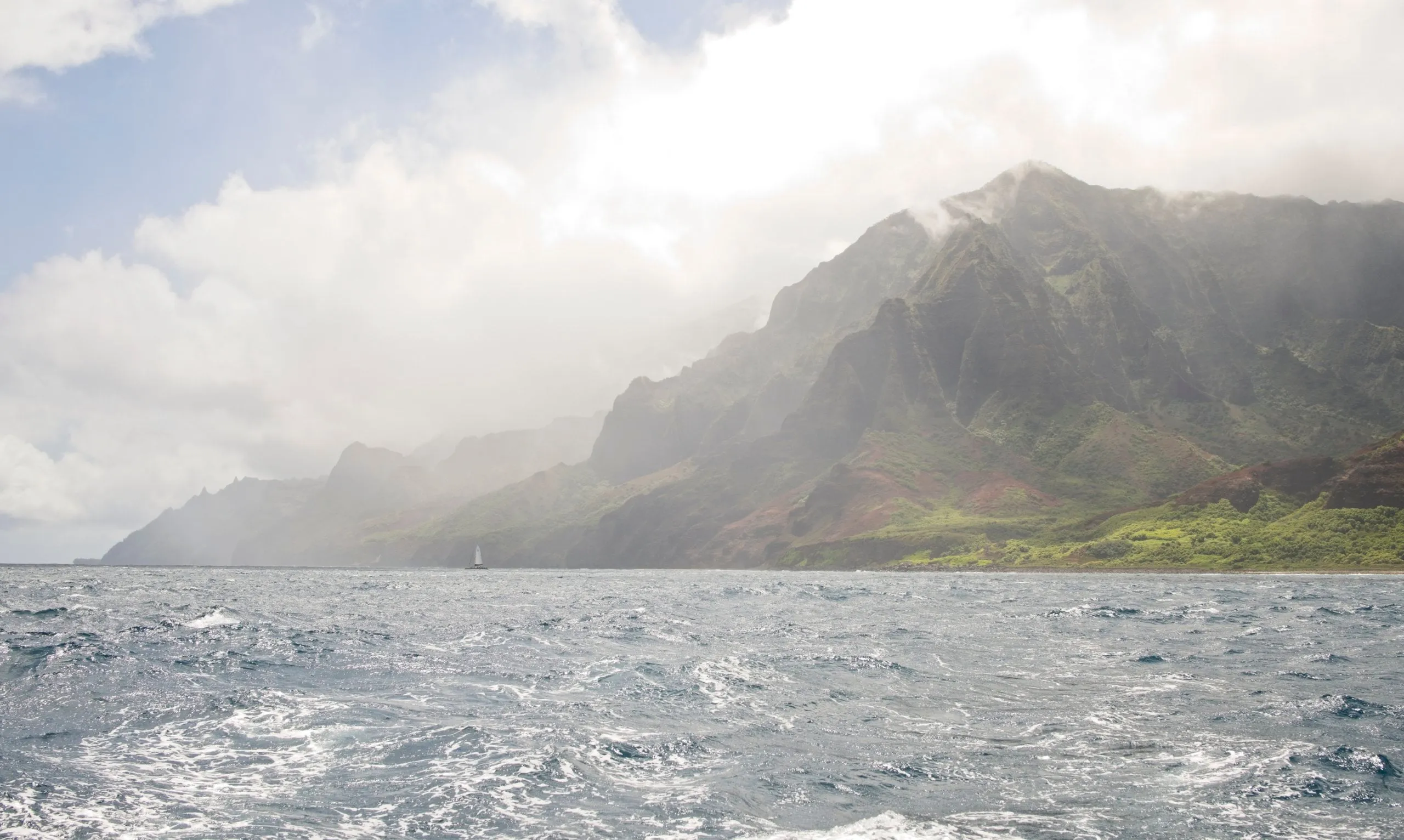 Sunlit haze drifting across the jagged cliffs of the Nā Pali Coast above a choppy blue sea, a small sailboat near the shore