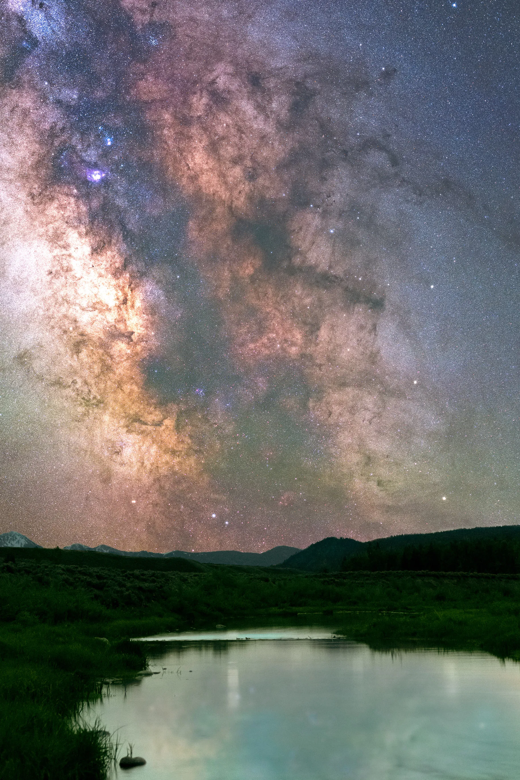 The Milky Way glowing above a green meadow and a calm stream reflecting the night sky