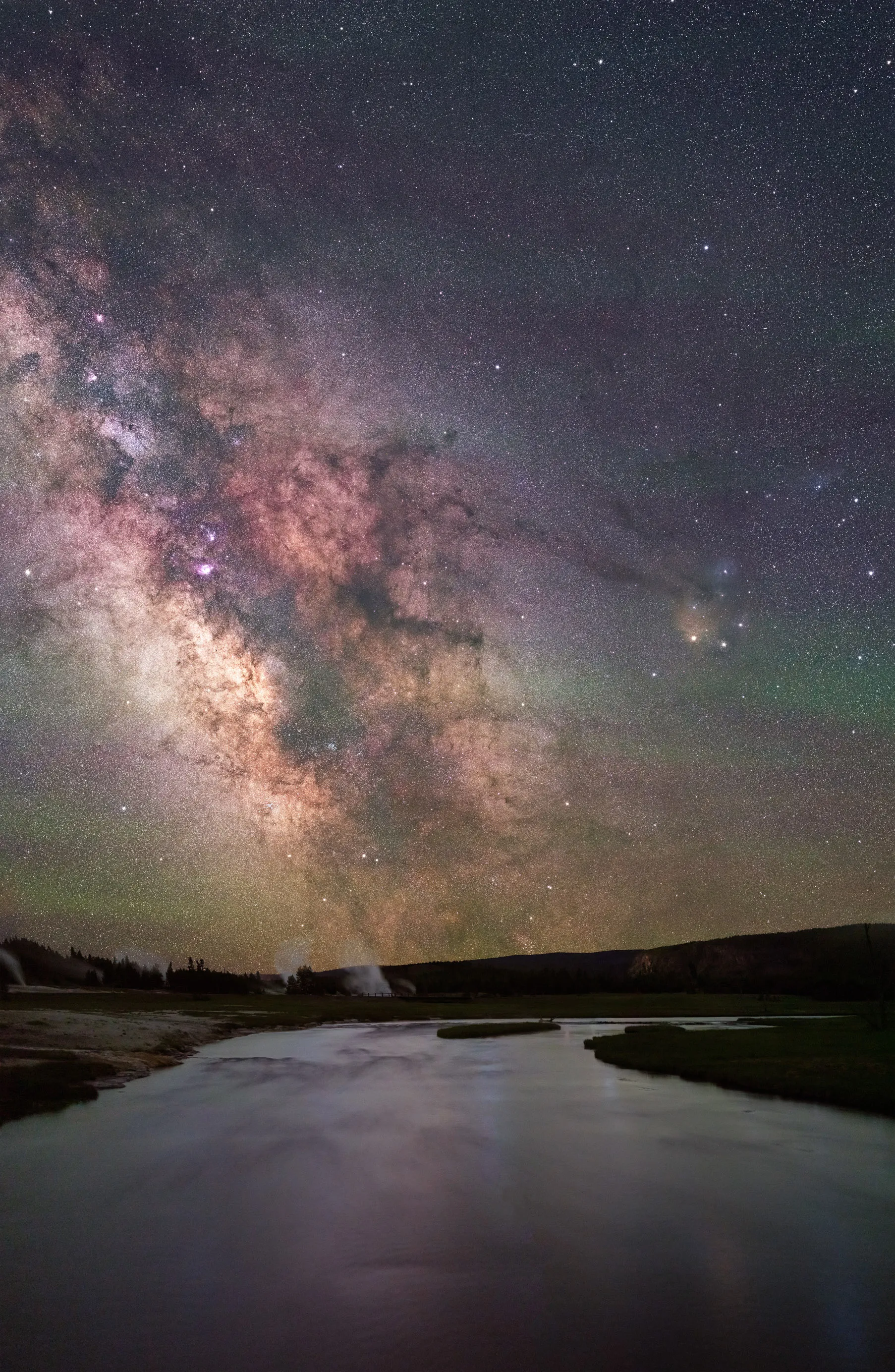 The Milky Way above a calm river, with steam rising from a distant geyser on the horizon