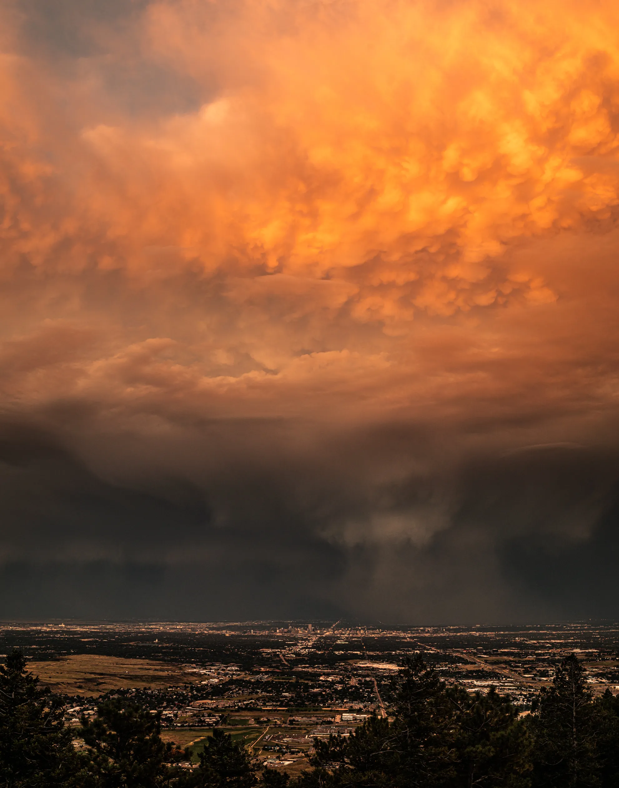 A fiery orange storm cloud above a dark curtain of rain and the lights of a city on the plains below