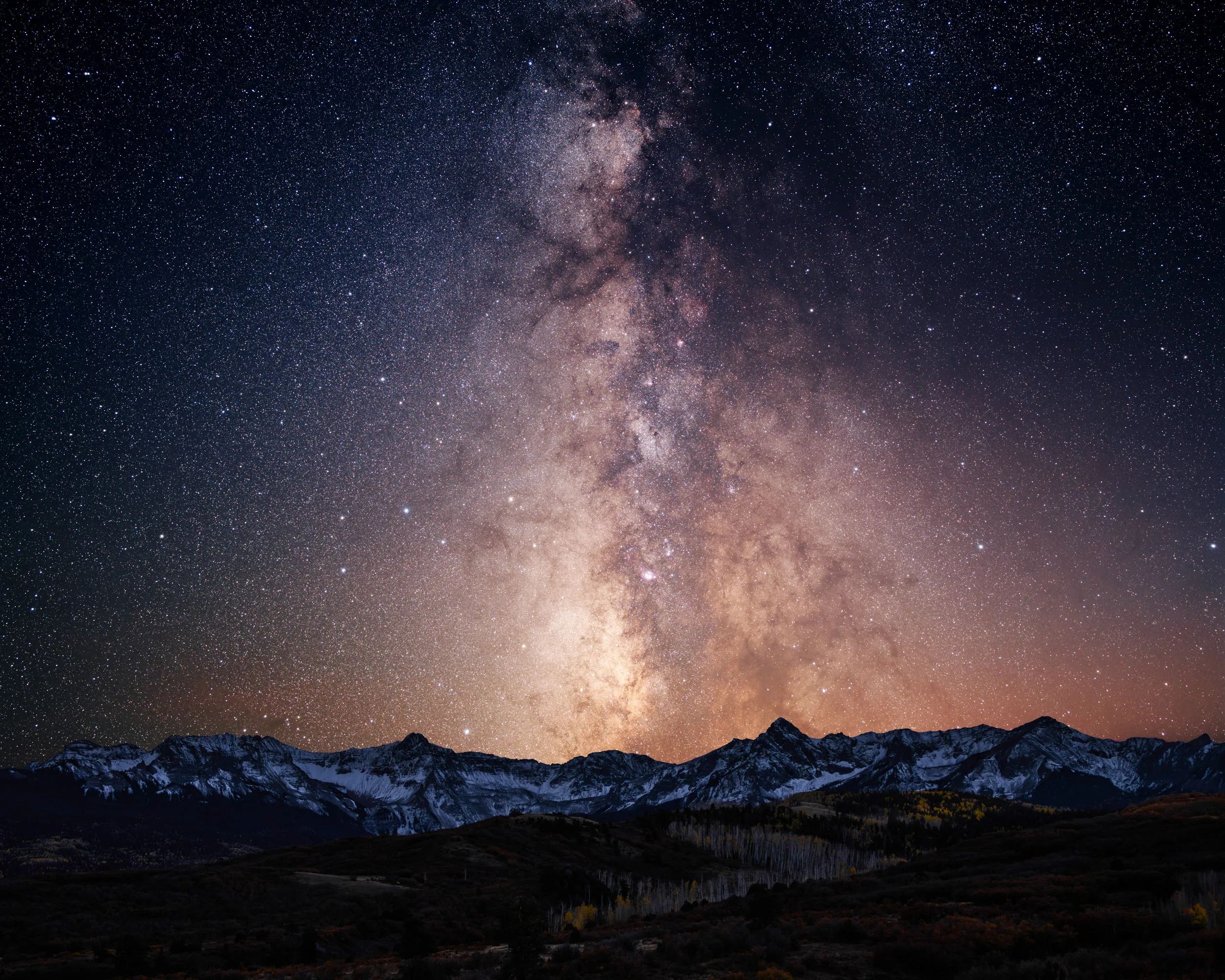 The Milky Way rising above a snowy mountain range, golden aspens faintly visible in the dark foreground
